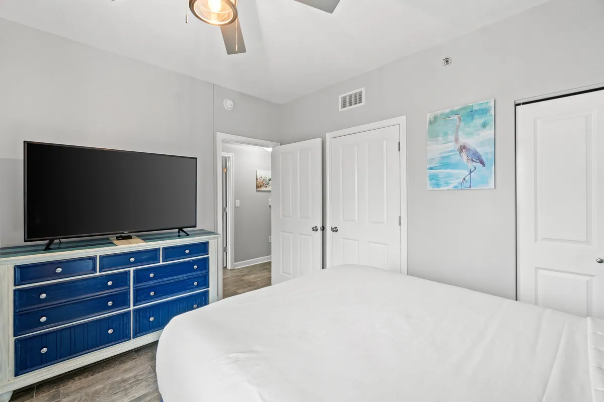 A serene bedroom with a blue dresser, large TV, tasteful artwork, and a bed with crisp white linens. The room is designed for rest and relaxation, offering guests a peaceful retreat after a day enjoying resort amenities.