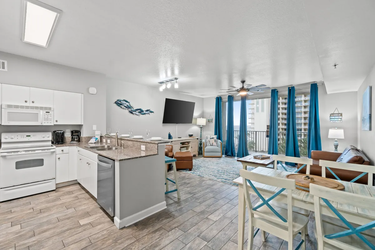 This image showcases the heart of the condo, featuring a stylish kitchen area, living room seating, and large balcony doors that open to the outside. Decorative wall art adds a touch of personality, and the space is bathed in natural light, emphasizing the open-concept layout. You can see both the kitchen and living areas as well as a glimpse of the inviting balcony beyond.