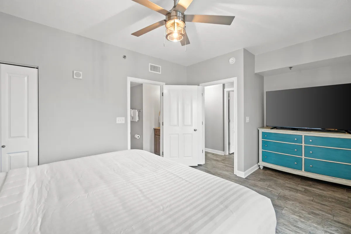 A cozy bedroom featuring a bed with crisp white linens, a blue dresser, and a ceiling fan overhead. The room is bathed in natural light, creating a soothing environment ideal for relaxation and rejuvenation.
