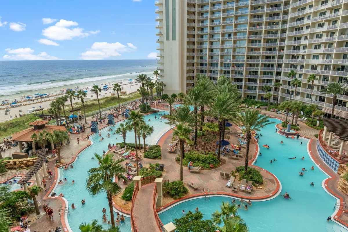 A vibrant outdoor pool area surrounded by tropical palm trees, with sunbathers lounging by the pool and the hotel building and beach just beyond. The pool area looks spacious and is perfect for group gatherings and family fun, with easy access to both the water and the sand.