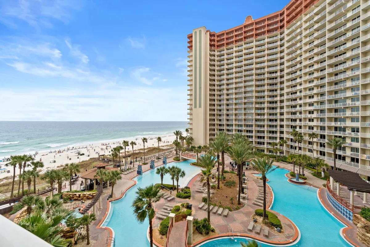 This exterior image features a stunning swimming pool surrounded by palm trees, with a sandy beach and a high-rise condominium in the background. Guests can be seen enjoying the pool and relaxing on sun loungers, all set against a vacation-like atmosphere.