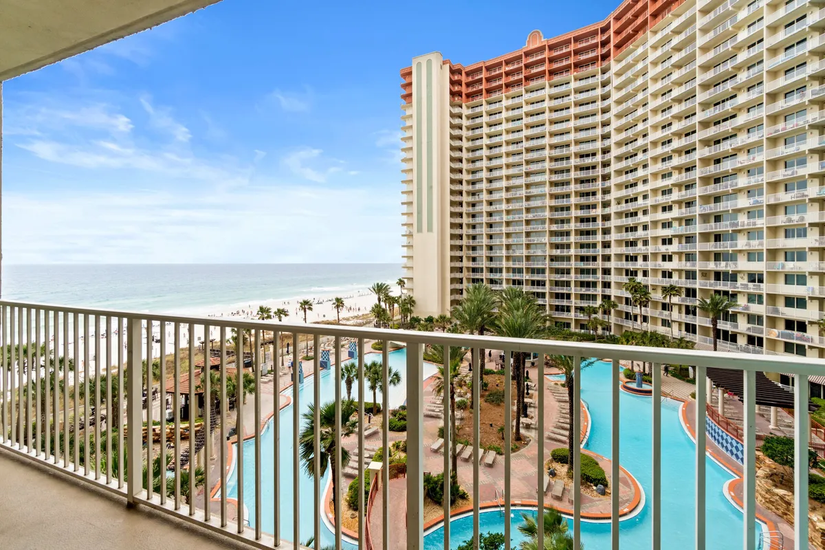 A sweeping exterior view of the resort, featuring a sparkling pool surrounded by palm trees, inviting lounge areas, and the striking architecture of the hotel with the Gulf of Mexico visible beyond. The scene captures both the pool area and the proximity to the ocean, evoking a luxurious vacation atmosphere.
