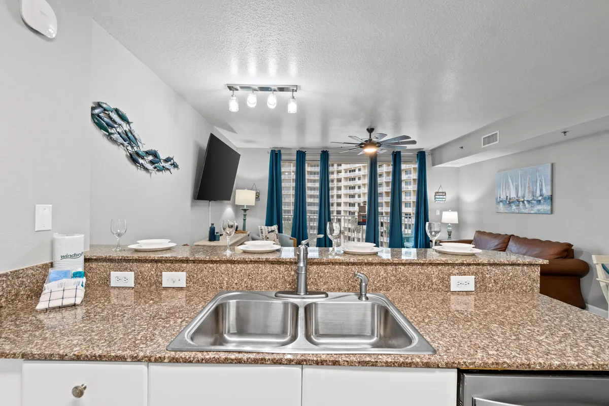 A view from the kitchen sink, looking out over granite countertops toward the living room seating area, blue curtains, and a television. Natural light fills the space, reinforcing the open, inviting layout where cooking and socializing blend seamlessly.
