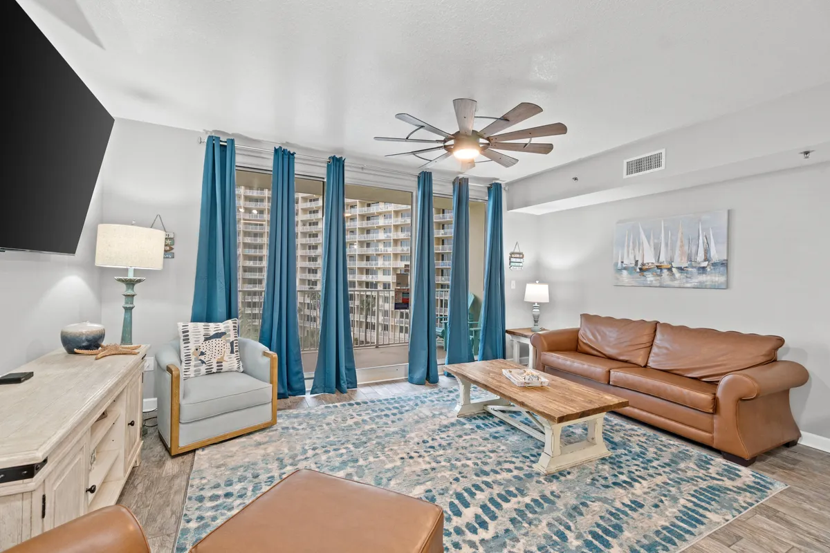 A cozy living room with a brown leather sofa, blue armchair, large patterned area rug, and floor-to-ceiling glass doors with blue curtains leading to a balcony. This is the main communal space, also accommodating the sleeper sofa mentioned in the section.