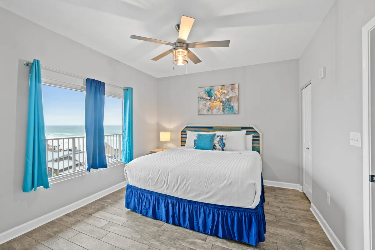 A serene master bedroom featuring a king-size bed with blue and white bedding, a large window offering an ocean view, a ceiling fan with a light fixture, and modern abstract wall art above the bed. The space is bright, airy, and inviting, with natural coastal light filling the room.