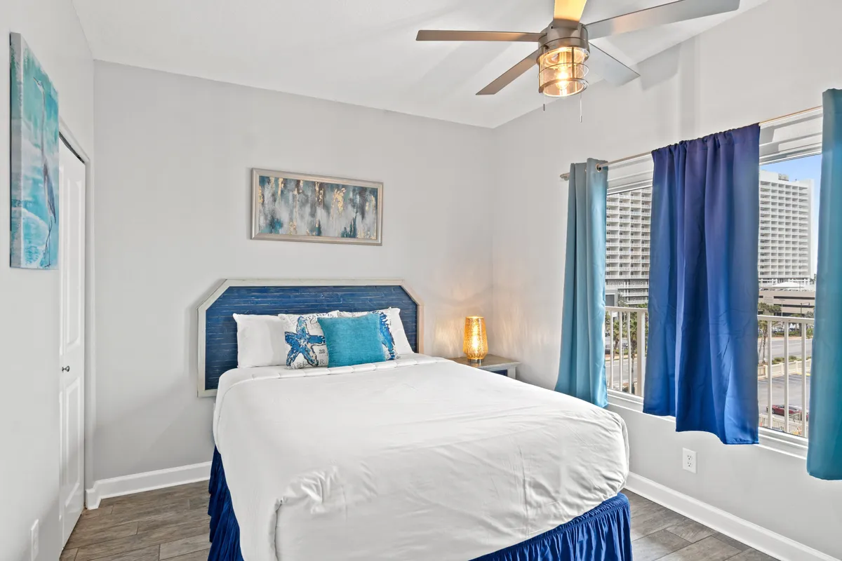 A calm and inviting second bedroom with a queen-sized bed dressed in blue and white bedding, a modern ceiling fan, a large window with blue curtains letting in natural light, and a small bedside table with a decorative lamp.