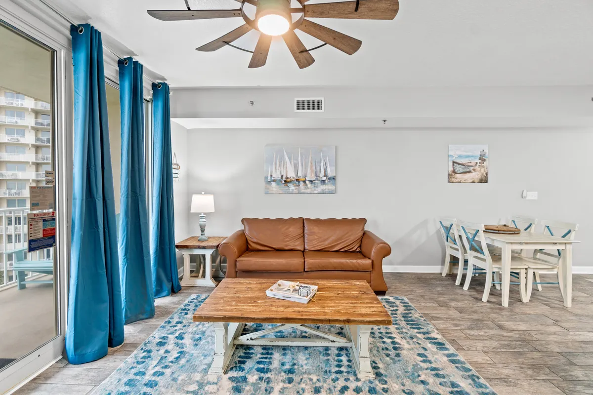 A spacious open-concept living and dining area furnished with a brown leather sofa, a wooden coffee table, a blue patterned area rug, and a dining table with chairs. Large sliding glass doors lead out to a balcony, providing an abundance of natural light and a direct view of the Gulf.