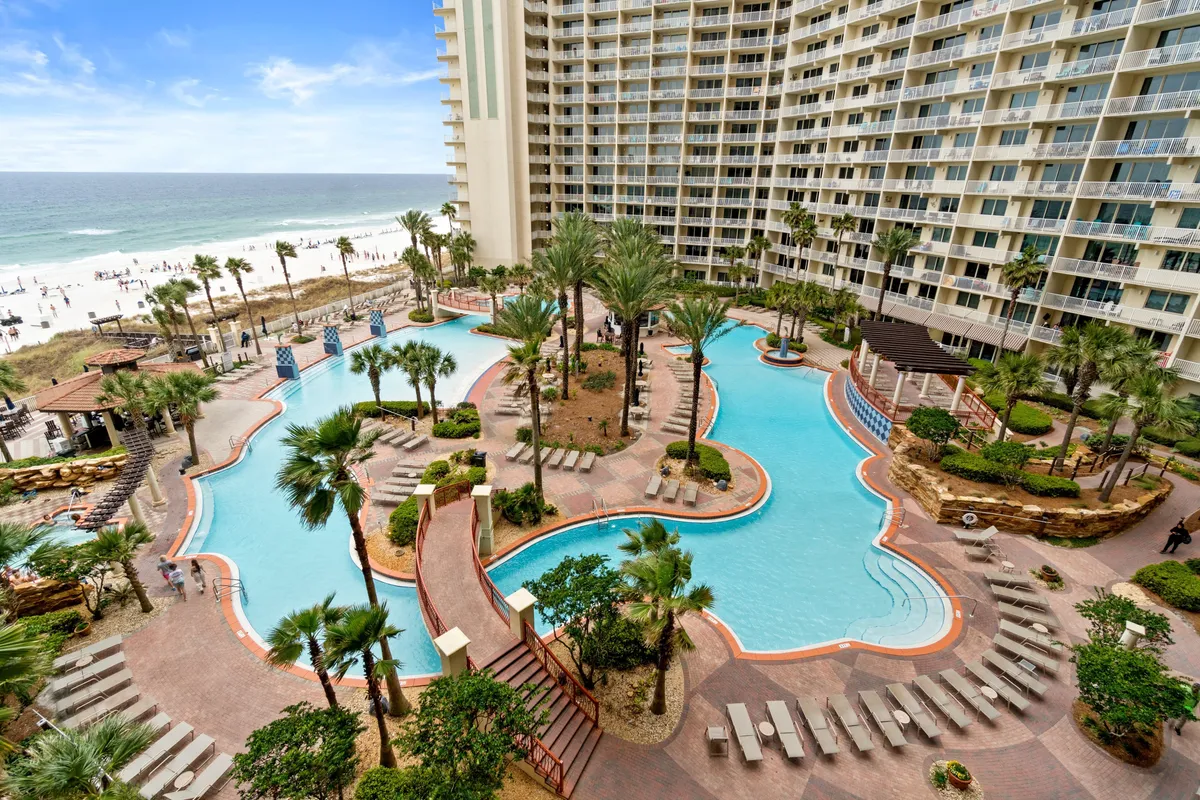 A sweeping view of the resort's lagoon-style swimming pool, complete with fountains, a charming bridge, tall resort buildings with balconies, and the Gulf of Mexico and sandy beach in the background. The image is lively, showing ample space for sunbathing, swimming, and socializing.