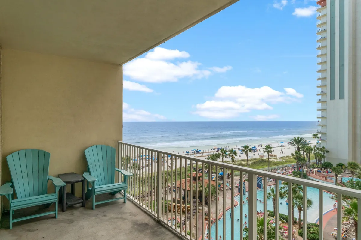 This image captures a private balcony with turquoise chairs overlooking the expansive Gulf of Mexico, the resort swimming pool, palm trees, and a lively beach filled with umbrellas and visitors. The scene effortlessly blends private relaxation with the vibrant life of the resort and the beach beyond.