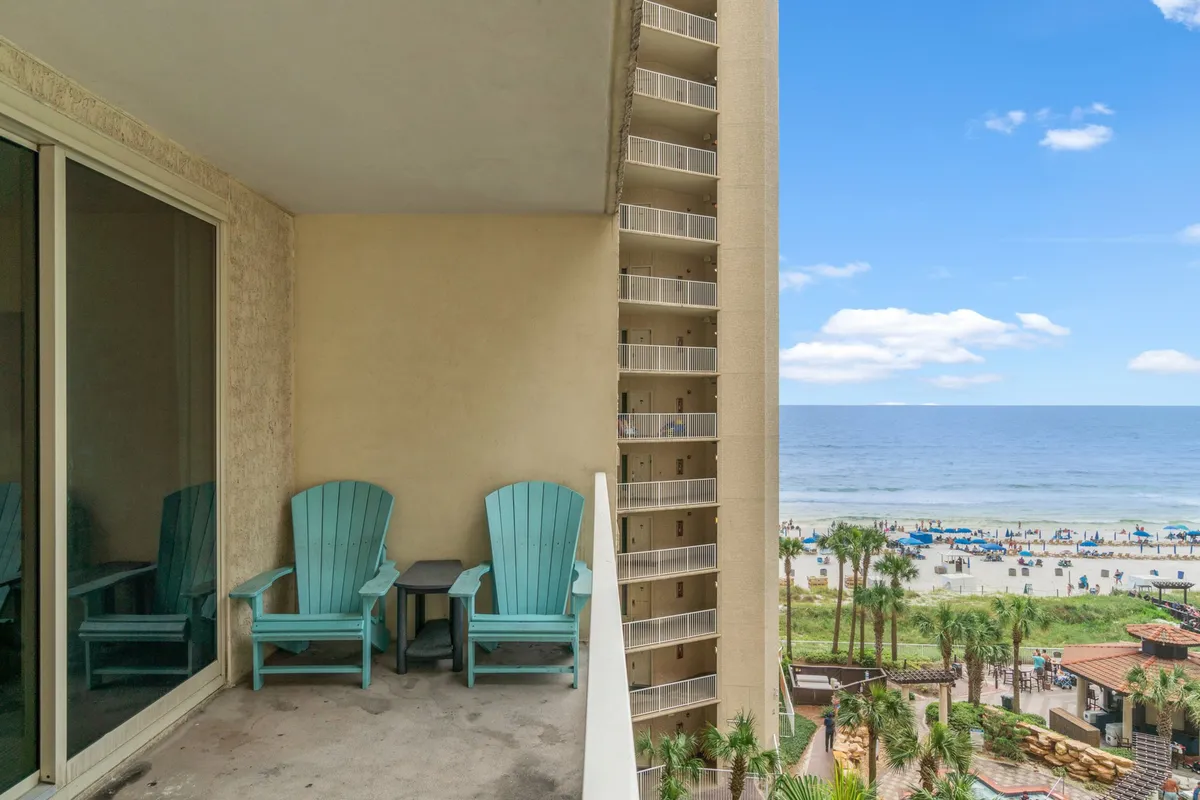 A private balcony with two turquoise Adirondack chairs overlooking a distant view of the beach, blue ocean, and sky. The setting is tranquil, with partial views of adjacent balconies and lively beach scenes below.