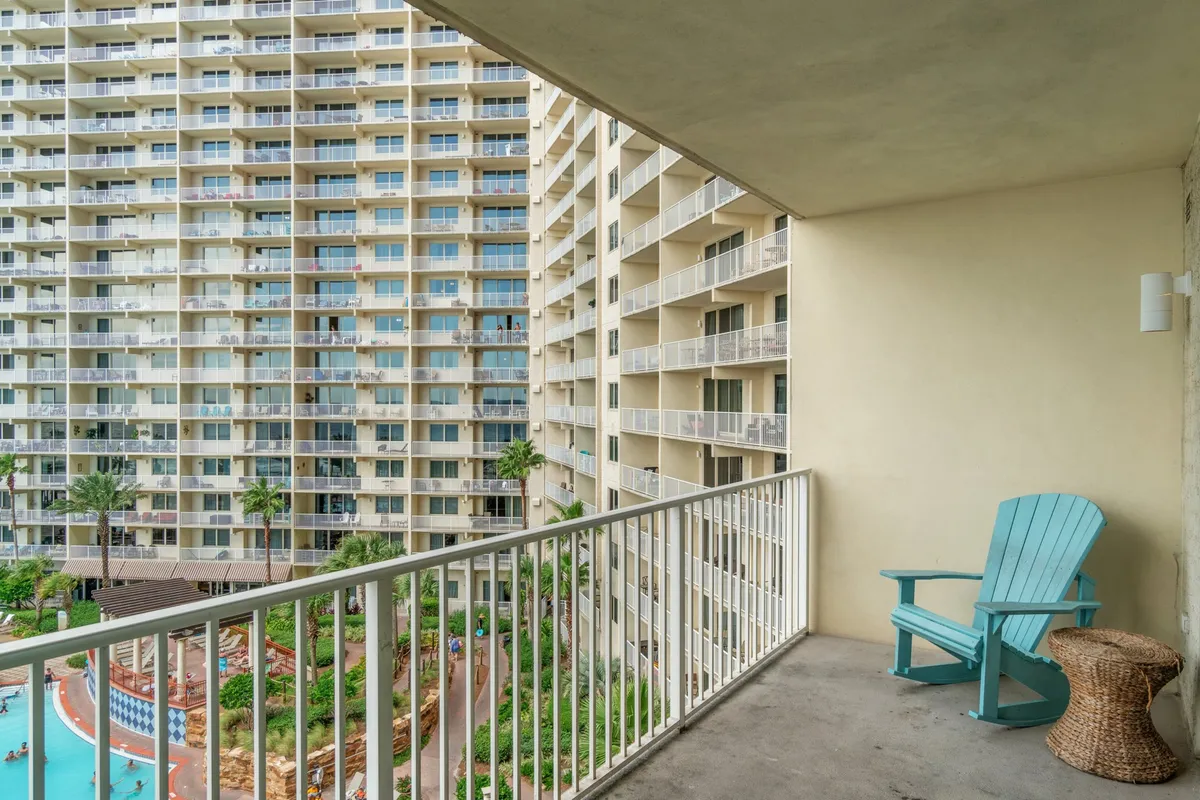 A bright turquoise Adirondack chair and wicker side table sit on a private balcony overlooking the resort pool and adjacent balconies, with a white railing framing the view. The image hints at both relaxation and connection to the vibrant resort life below.