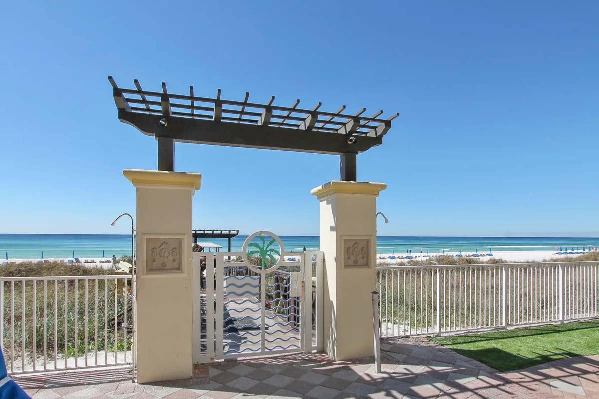 A private balcony with a decorative pergola frames breathtaking panoramic views of the turquoise Gulf waters and pristine white sandy beach. The gated entrance hints at exclusive, direct access to the beach below, with a white railing offering both safety and unobstructed sightlines to the ocean horizon.