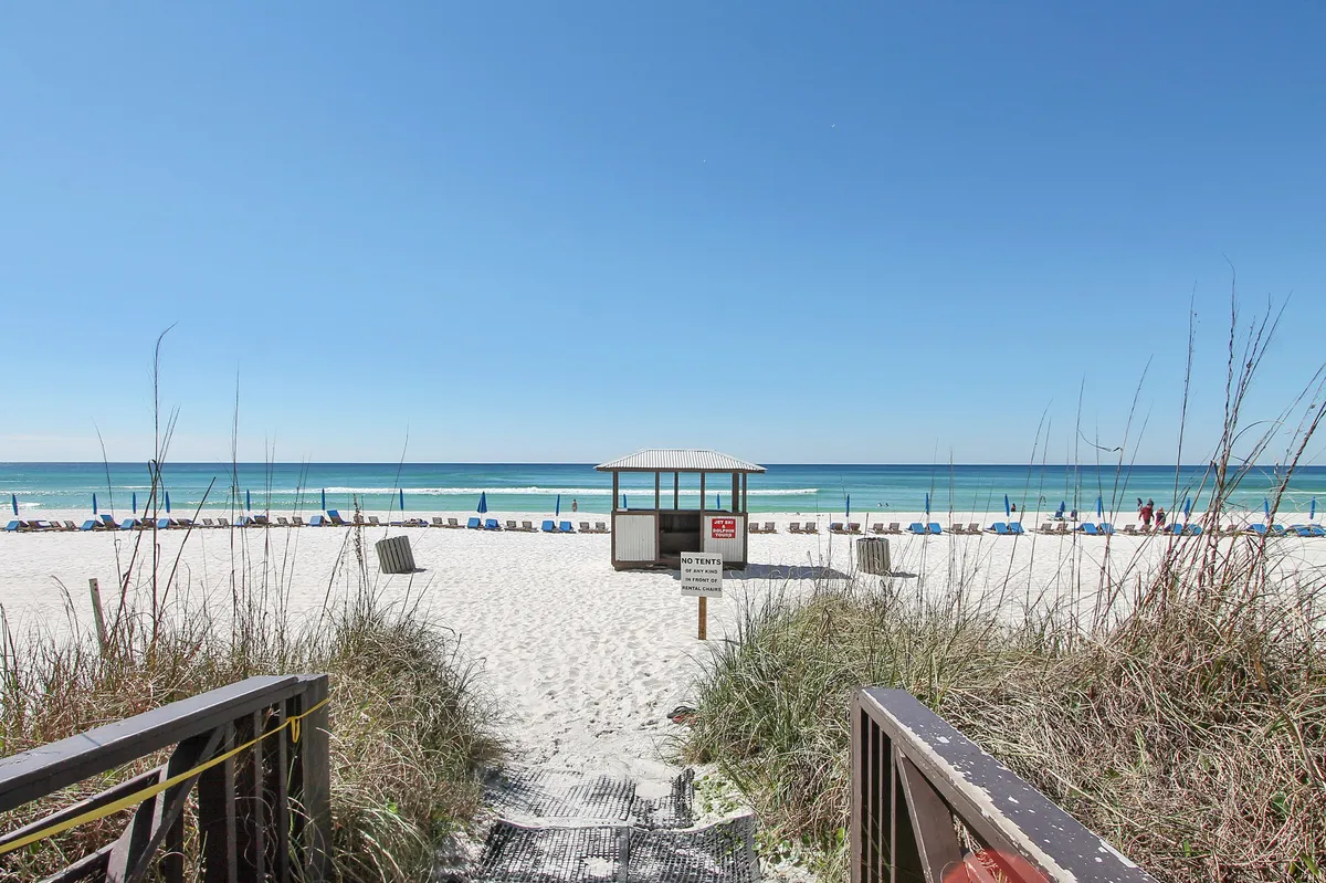A broad pathway winds through dune grass toward a classic lifeguard hut on a pristine white sand beach, with rows of blue umbrellas and chairs set along the shore. The calm emerald-green Gulf waters stretch out under a clear sky, evoking the excitement of stepping straight onto the beach from the resort.