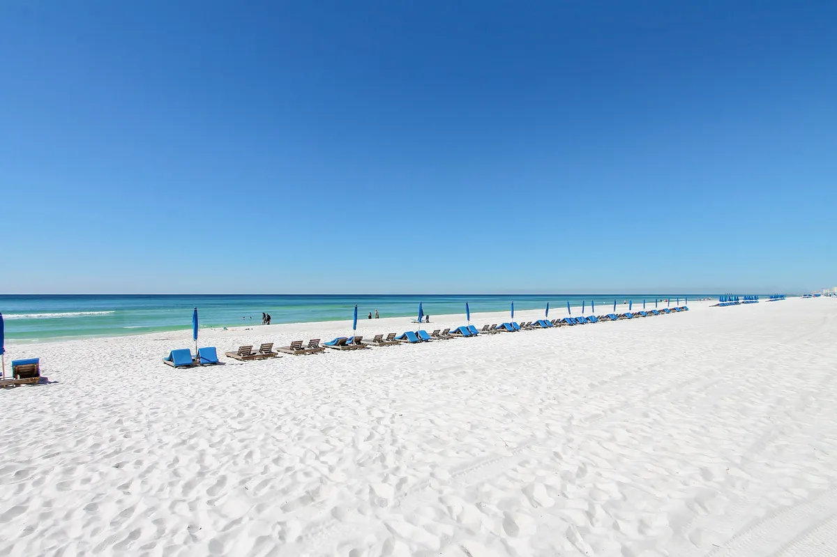 A stretch of pristine white sandy beach adorned with neatly arranged blue lounge chairs and umbrellas faces the calm, turquoise waters of the Gulf. The horizon is clear, offering an uninterrupted view for sunset watching and tranquil beach strolls.
