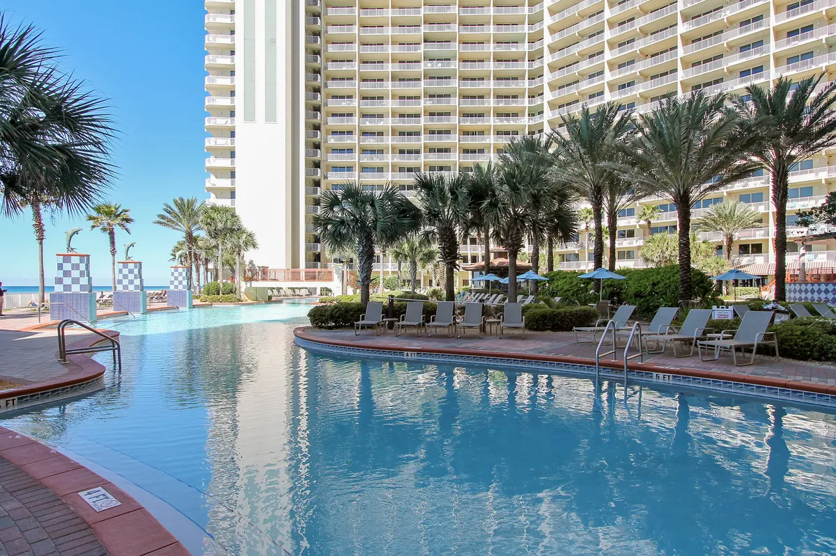 A sprawling lagoon-style swimming pool with elegant fountains sits at the heart of the resort, surrounded by tall palm trees, sun loungers, and umbrellas. The multi-story resort building with balconies rises in the background, evoking the grandeur and tropical ambiance of a high-end beachside retreat.