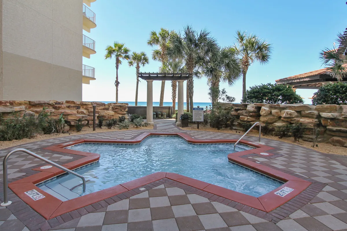 A star-shaped hot tub bubbles invitingly near palm trees and stone walls, with a pathway leading towards a decorative pergola and beach access. The scene exudes relaxation and easy movement from poolside leisure to the sandy shore.