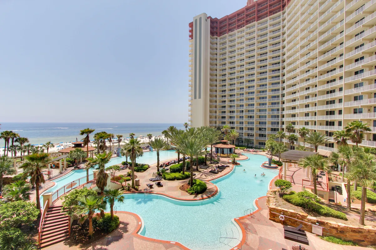 A large lagoon-style swimming pool with playful fountains is surrounded by lush palm trees and a multi-story resort building with balconies. The scene opens to a private beach with the ocean in the background. The lively pool area also hints at nearby social amenities, such as the tiki bar, and offers easy access to both pool and beach activities.