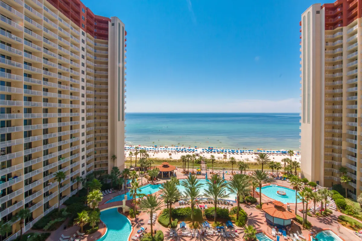 This vibrant exterior shot captures two tall condominium towers framing a large lagoon-style swimming pool, with palm trees lining the resort grounds. The private beach, with rows of umbrellas and lounge chairs, stretches out to the calm Gulf of Mexico waters under a blue sky.
