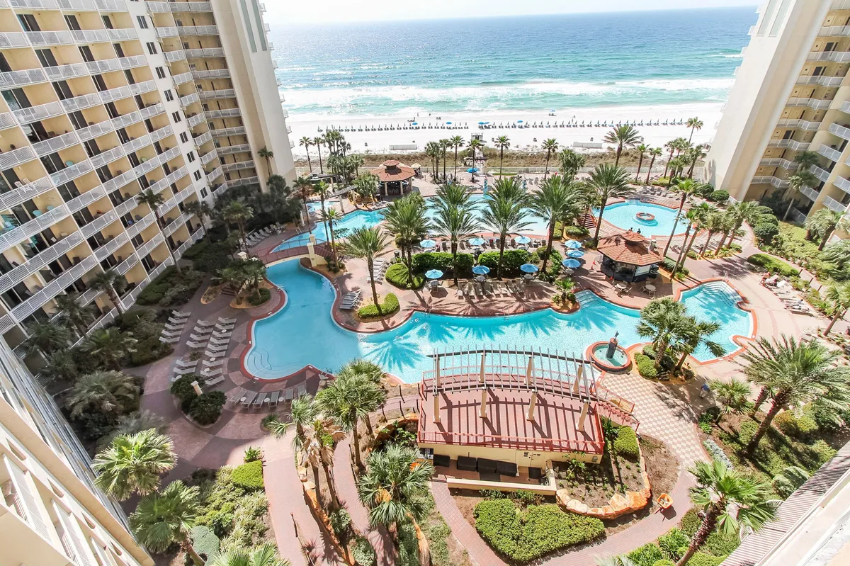 This exterior image captures a large lagoon-style swimming pool surrounded by palm trees, with two tall resort buildings framing the pool area. The private beach with white sand and Gulf of Mexico waves can be seen in the background, reinforcing the sunny, resort-like atmosphere.