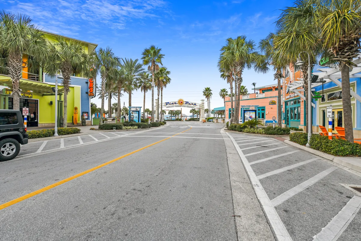 A sunny exterior view of Pier Park’s colorful storefronts, palm-lined street, and the iconic archway sign, with an open road leading towards the beach. The inviting environment suggests plenty of options for dining, shopping, and meeting up with friends.