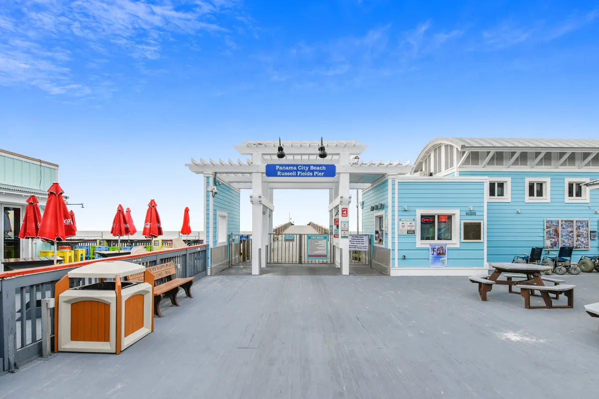 This exterior scene shows the entrance archway to the Panama City Beach Russell Fields Pier, with a blue building, red umbrellas, picnic tables, and benches. The area is lively and inviting, encouraging exploration of local attractions and community.