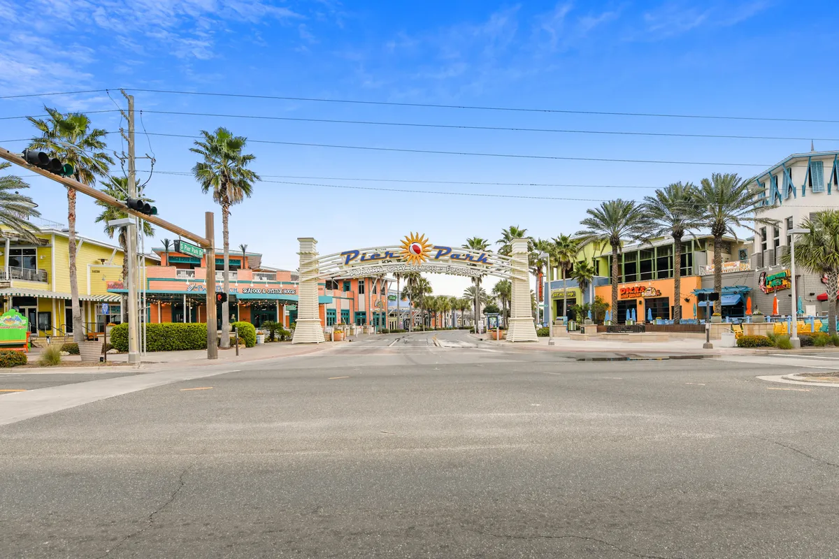 The entrance archway to Pier Park is featured, with colorful storefronts, palm-lined streets, and a clear blue sky. The scene is bustling with energy, suggesting shopping, dining, and group-friendly entertainment options within walking distance of the resort.