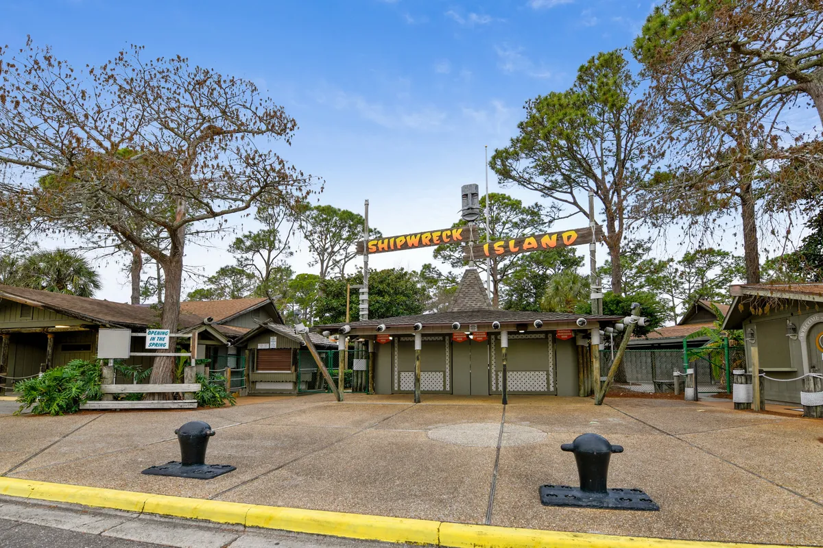 The entrance to Shipwreck Island Water Park, with its playful themed sign and rustic buildings, invites families to a day of adventure and water fun. The open plaza and surrounding trees create a welcoming, festive atmosphere, signaling one of the top complimentary attractions available to guests.